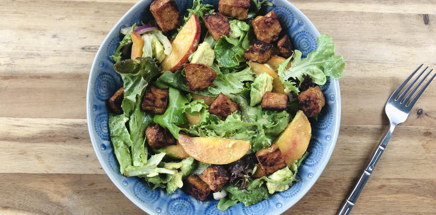 vibrant green salad with peach slices and tempeh in a blue bowl on a wooden surface