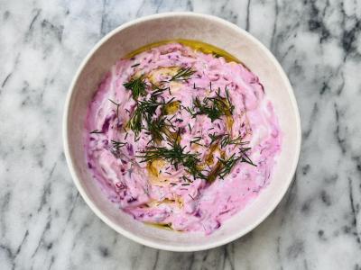 a bowl of magenta hued dip garnished with dill and olive oil in a white bowl atop a marble surface