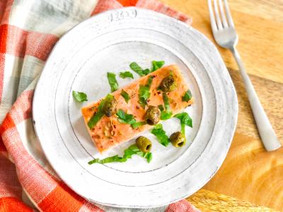 a portion of cooked salmon with olives and herbs on a white plate resting on a wooden table