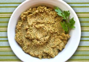 Creamy pesto hummus in a bowl, topped with olive oil and herbs, accompanied by colorful vegetable sticks and pita chips.