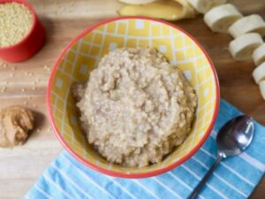 A colorful bowl filled with creamy oatmeal, surrounded by sliced bananas, peanut butter, and millet on a wooden surface.