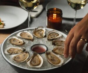 A hand reaching for an oyster from a circular platter of twelve oysters on ice with wine glasses nearby.