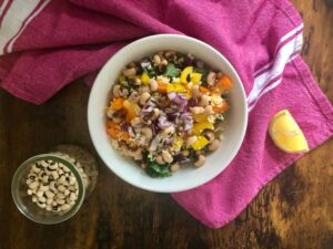A colorful salad with black-eyed peas, red onion, yellow bell pepper, kale, quinoa, and lemon wedge on a pink cloth.