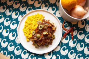 A plate of yellow rice with meat topped with olives, set on a tablecloth with a swan pattern.