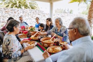 Family enjoying a meal together outdoors with traditional dishes and colorful tablecloth.