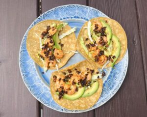 Three shrimp tacos with avocado and cabbage on a blue plate.